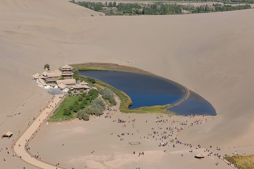 Le lac du Croissant vu du haut des dunes : un plan d'eau en forme de faucille bordé de roseaux et d'un bosquet vert, un ensemble de pavillons de bois à toits recourbés sur sa rive, et tout autour la masse ocre des dunes du mont Mingsha parcourue de visiteurs
