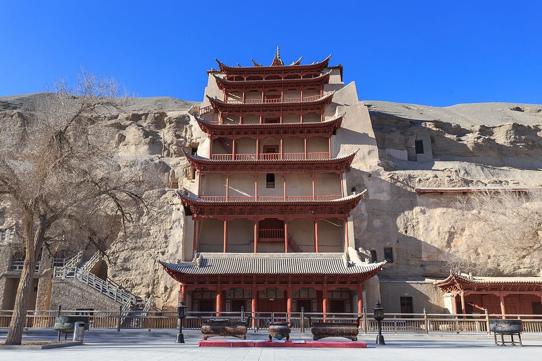 Le pavillon à neuf étages des grottes de Mogao : une structure de bois peinte en rouge vif adossée à une falaise de roche claire, ses toits recourbés superposés, et à droite dans la paroi plusieurs ouvertures de grottes protégées par des portes