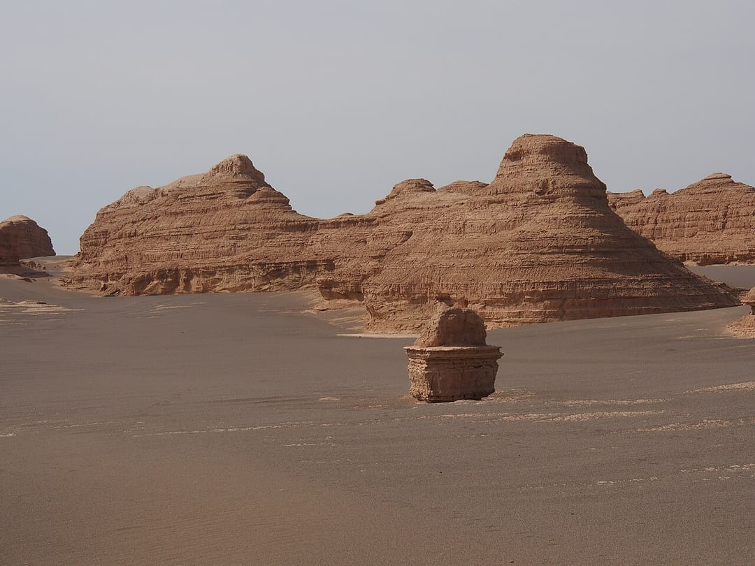 Formations rocheuses du Yadan : plusieurs buttes de roche ocre aux strates horizontales marquées, érodées en formes arrondies et allongées, posées sur une étendue de sable plat et lisse sous un ciel pâle