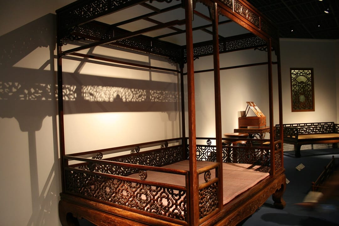 Lit à baldaquin chinois d'époque Ming en bois sombre : quatre montants sculptés portant un dais ajouré, galeries de treillis fermant trois côtés de la couche, exposé dans une salle de musée