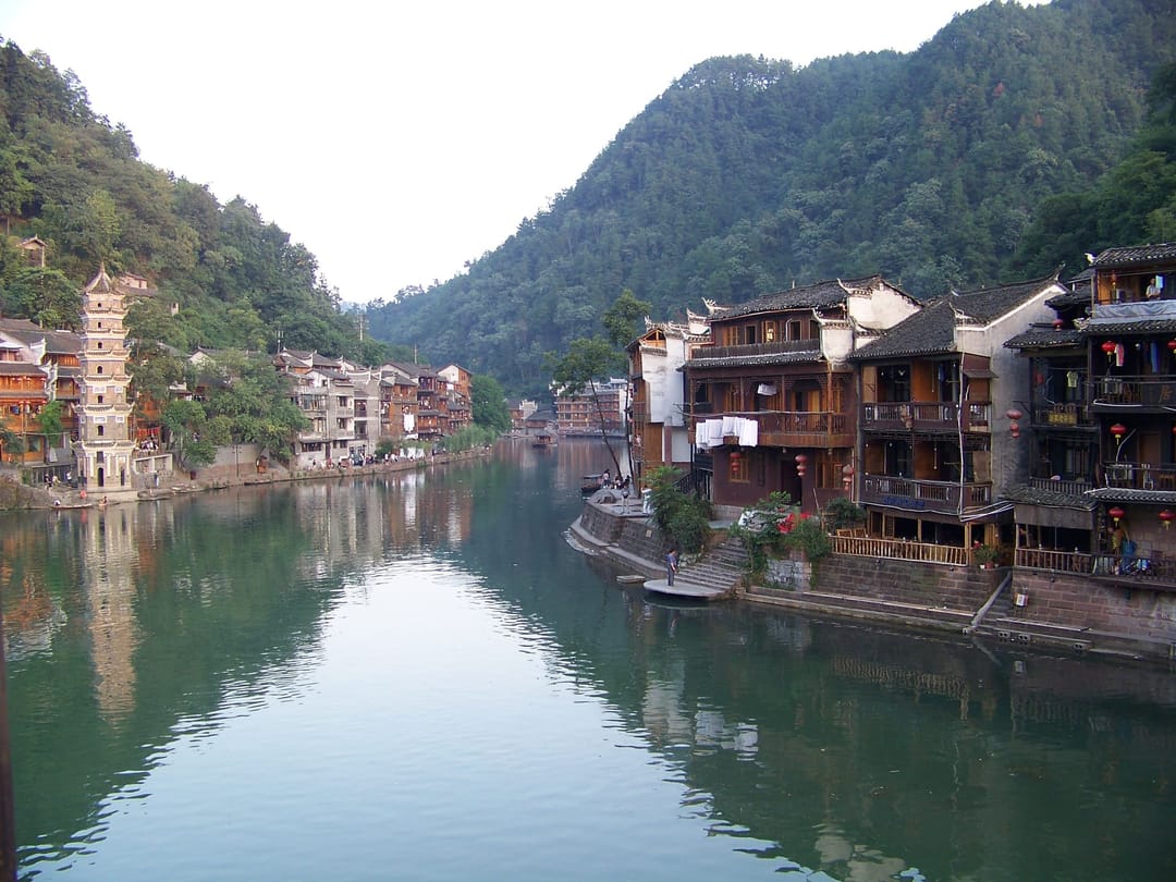 La rivière Tuo à Fenghuang vue depuis un pont : maisons de bois à balcons bâties au ras de l'eau sur la rive droite, lanternes rouges accrochées aux façades, pagode blanche à étages sur la rive opposée, collines boisées au fond et une barque amarrée