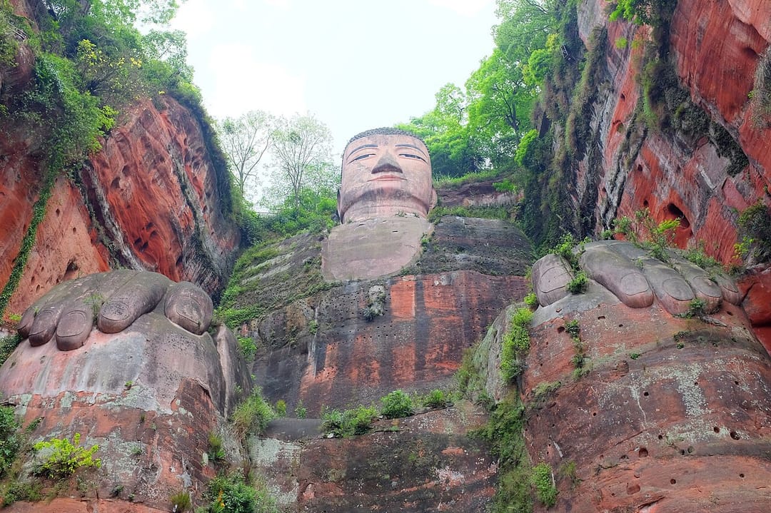 Le grand Bouddha de Leshan vu depuis ses pieds : deux pieds de pierre énormes occupent tout le premier plan, le corps s'élève derrière eux dans la falaise de grès rouge, et la tête apparaît toute petite au sommet contre un ciel pâle
