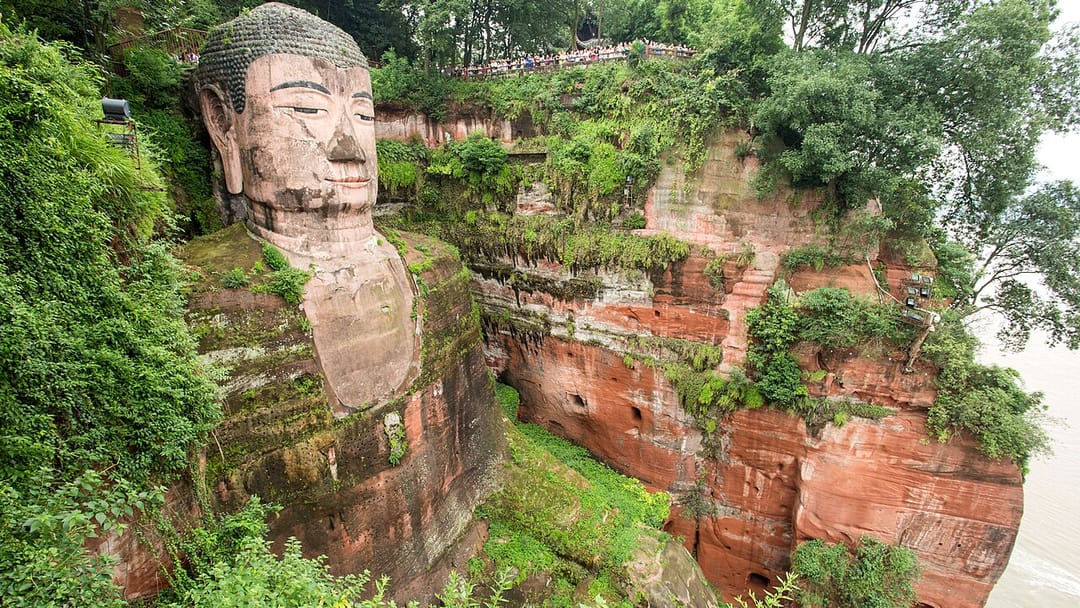 Le grand Bouddha de Leshan vu de côté depuis la plateforme supérieure : la tête et le buste taillés dans une falaise de grès rouge envahie de végétation, une file de visiteurs alignée sur la terrasse au-dessus de sa tête, et le fleuve visible à droite