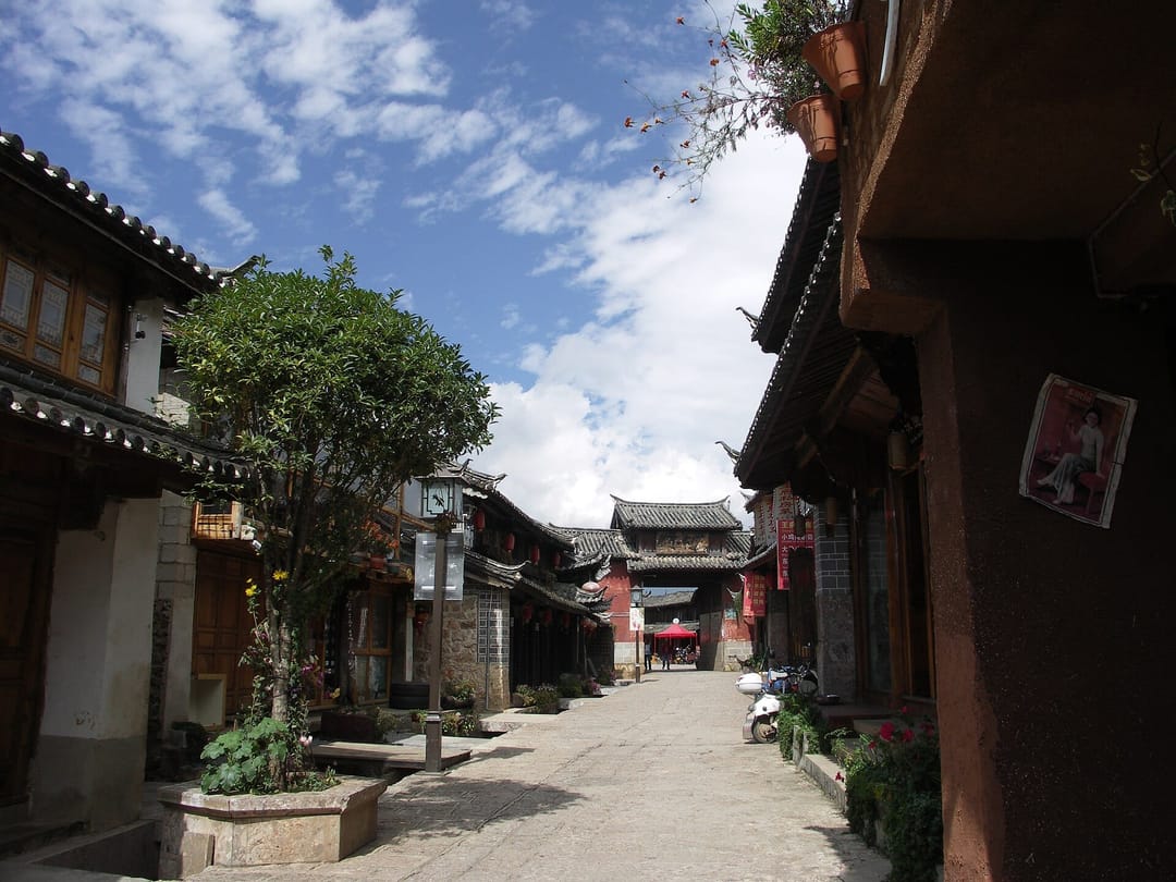 Rue pavée déserte de Baisha bordée de maisons naxi en bois et pisé aux toits de tuiles grises, avec une porte ancienne peinte en rouge au fond de la perspective