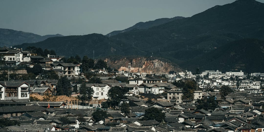 La vieille ville de Lijiang vue de hauteur : au premier plan une masse serrée de toits de tuiles grises aux faîtages recourbés, quelques façades blanches et des arbres entre eux, et derrière des crêtes montagneuses boisées qui ferment la vallée