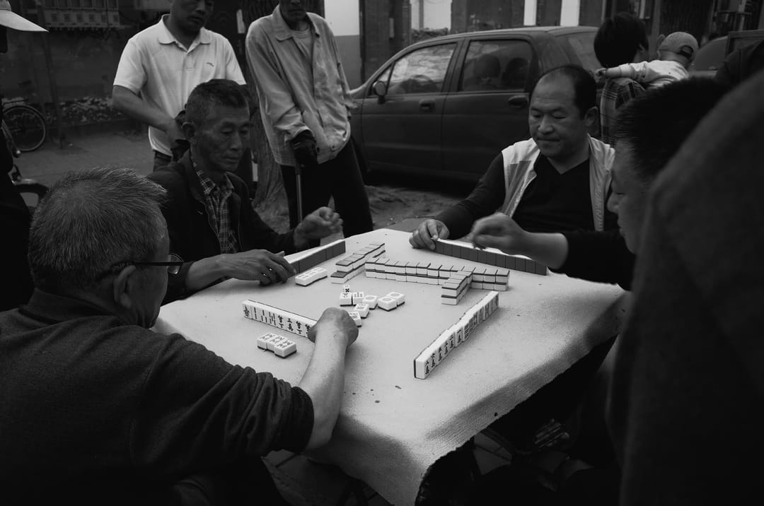 Partie de mahjong dans une ruelle de Pékin : quatre joueurs autour d'une table pliante couverte d'un drap, murailles de tuiles dressées devant chacun, des passants regardent par-dessus leurs épaules