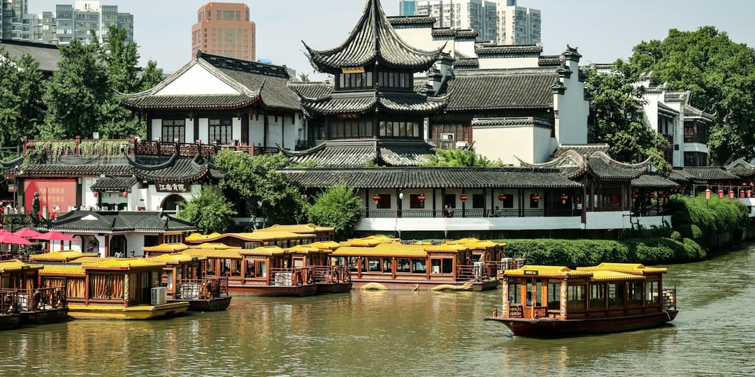 Le quartier du temple de Confucius à Nankin vu depuis la rivière Qinhuai : pavillons blancs à toits de tuiles grises recourbées et lanternes rouges alignées sous les auvents, une rangée de bateaux de promenade à toit jaune amarrés au premier plan, et des tours d'habitation modernes en arrière-plan