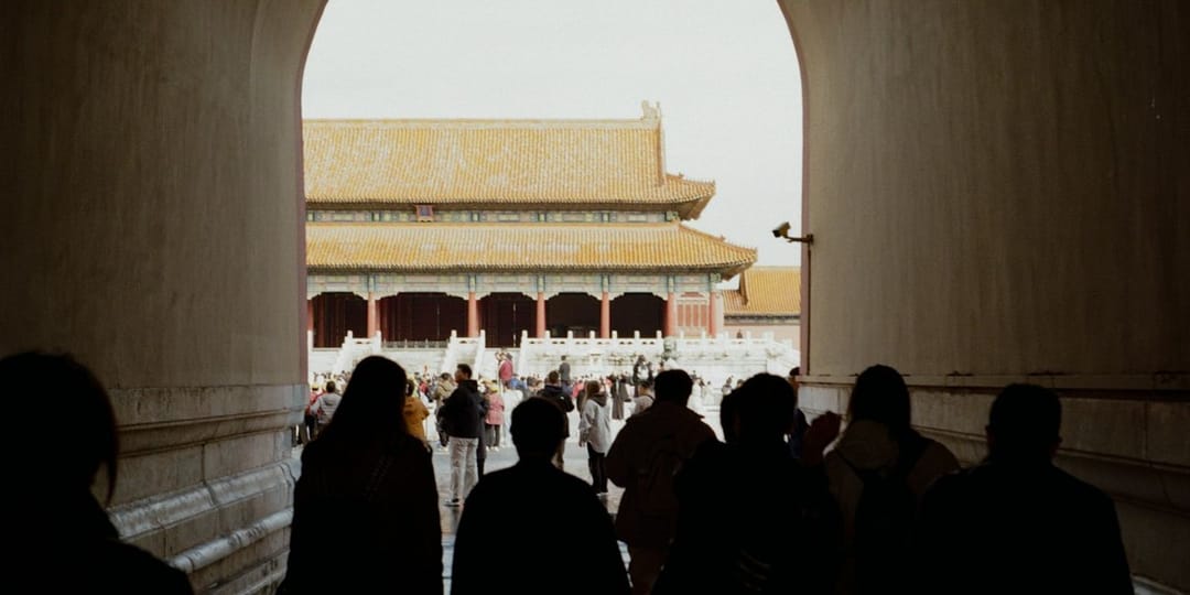 Vue depuis l'intérieur d'un passage voûté de la Cité interdite : des visiteurs en contre-jour avancent vers la sortie, et au-delà de l'arche s'ouvre une cour où se dresse un pavillon impérial à toit de tuiles jaunes, entouré d'une foule