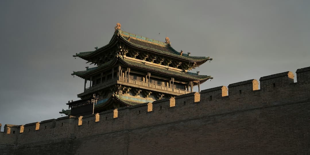 Une tour de porte à trois niveaux de toits de tuiles vernissées vertes se dresse au-dessus du rempart de brique crénelé de Pingyao, éclairée en lumière rasante dorée sur un ciel gris d'orage