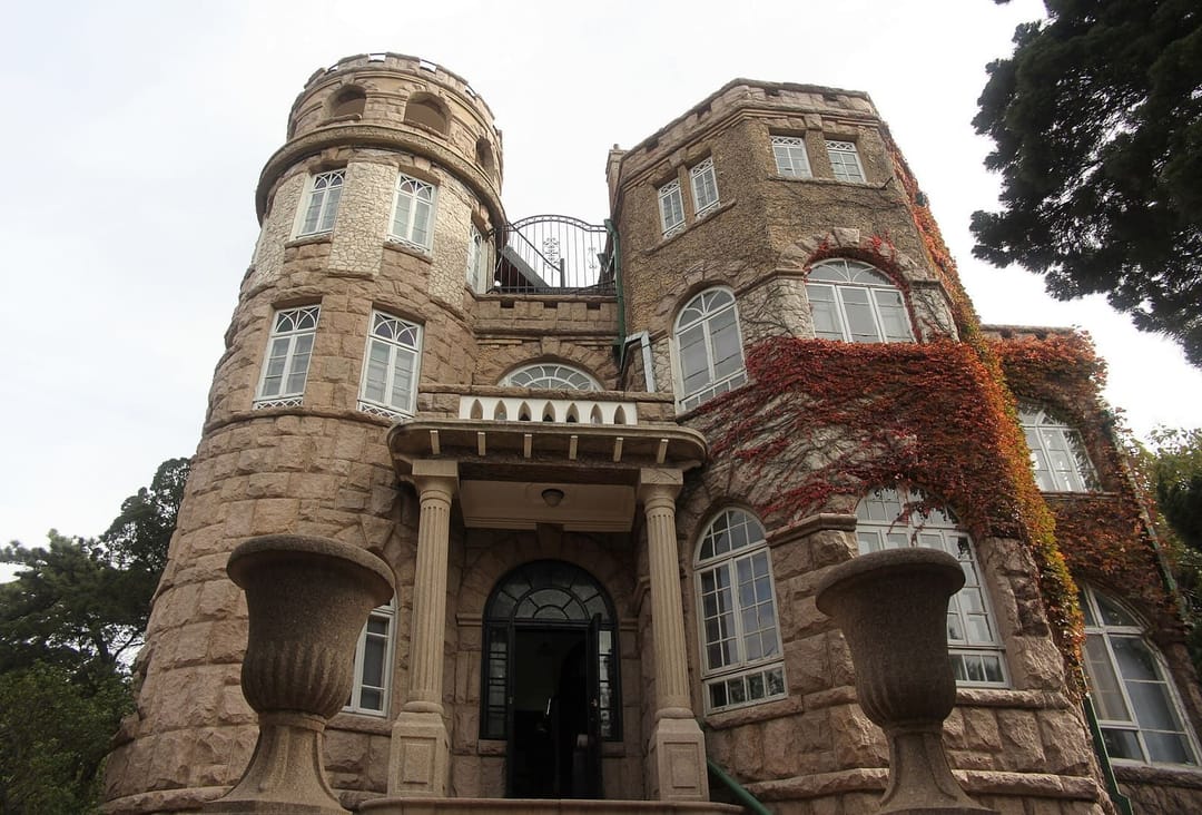 La villa Huashilou à Badaguan, Qingdao, vue en contre-plongée : une bâtisse de granit rose à tour ronde crénelée, fenêtres à petits carreaux blancs, porche à colonnes, et une vigne vierge rouge et orange qui couvre une partie de la façade