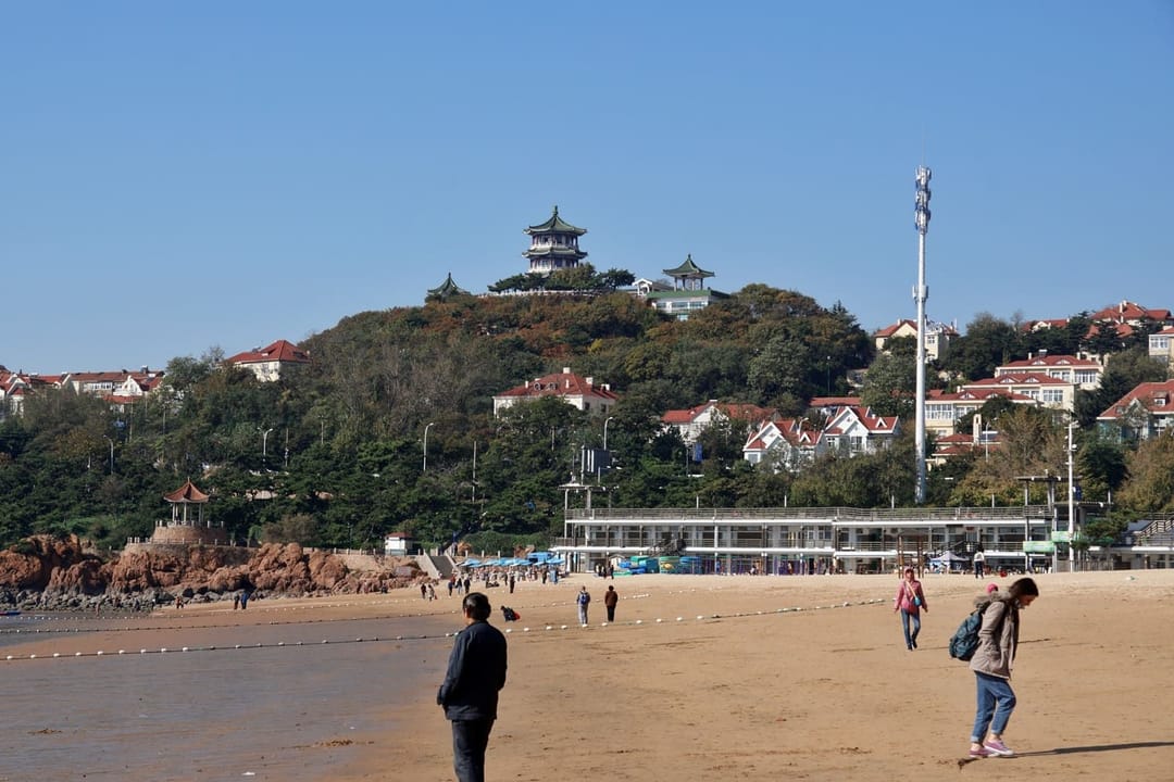 La première plage de Qingdao hors saison : une grande étendue de sable presque vide, quelques promeneurs en manteau, la ligne de bouées encore en place le long de l'eau, et sur la colline derrière les villas à toits rouges de Badaguan surmontées d'un pavillon chinois