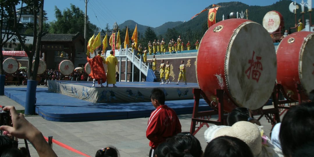 Une démonstration d'arts martiaux au temple de Shaolin : sur un tapis bleu surélevé, des pratiquants en robes jaunes et orange évoluent devant des rangées d'élèves alignés en tenue jaune, avec de grands tambours rouges au premier plan, des bannières jaunes, un public dense qui photographie, et les collines boisées du mont Song à l'arrière-plan