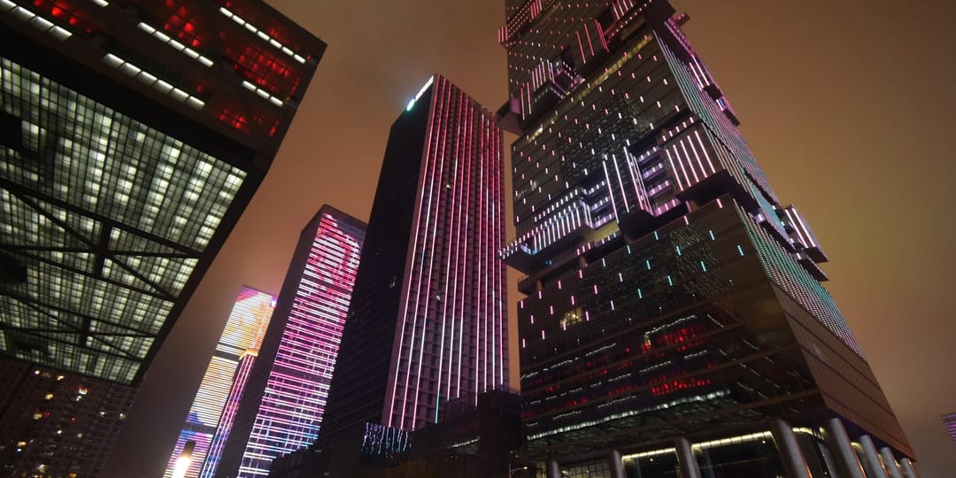 Les tours du quartier d'affaires de Futian à Shenzhen photographiées de nuit en contre-plongée : leurs façades sont couvertes de diodes qui dessinent des motifs roses, bleus et blancs synchronisés d'un immeuble à l'autre, sous un ciel orangé par les lumières de la ville
