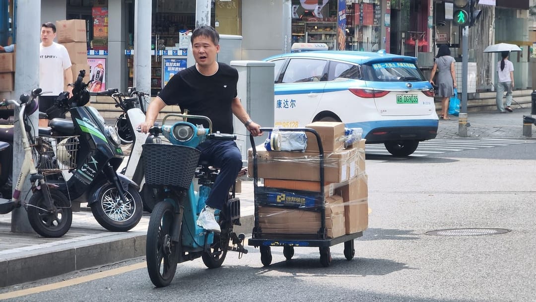 Un homme sur un deux-roues électrique tire un diable chargé de cartons scotchés dans une rue de Huaqiangbei à Shenzhen, avec un taxi bleu et blanc à plaque verte arrêté au feu derrière lui