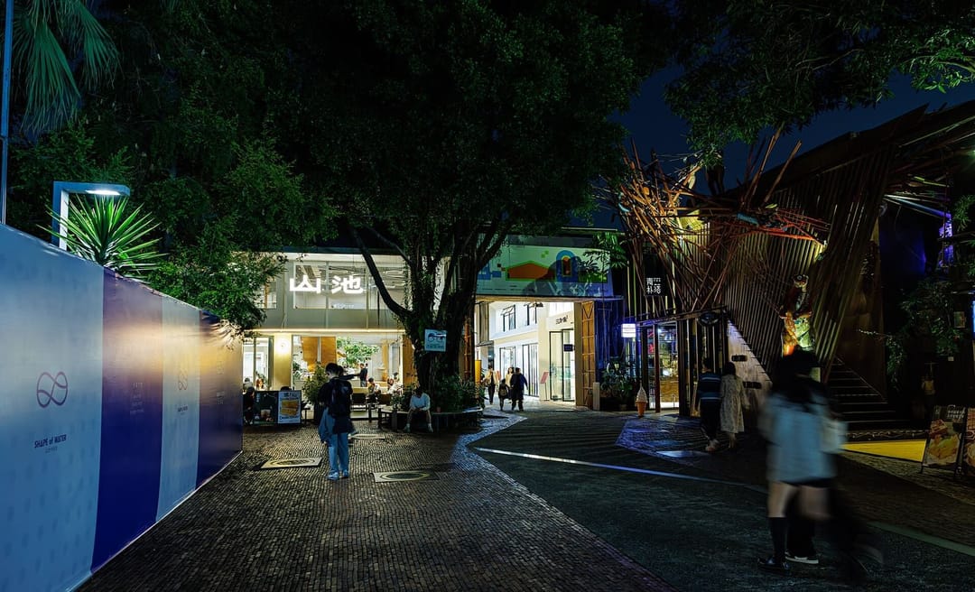 Une allée pavée d'OCT Loft à Shenzhen la nuit : d'anciens bâtiments industriels convertis en cafés et boutiques éclairés, de grands arbres au-dessus de la ruelle, une installation en bambou sur la droite et quelques passants