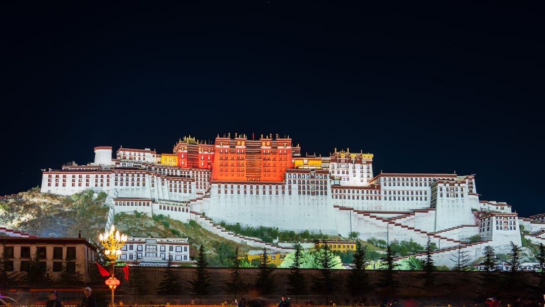 Le palais du Potala illuminé la nuit à Lhassa, façades blanches et corps central rouge étagés sur la colline, escaliers en zigzag et sapins au premier plan