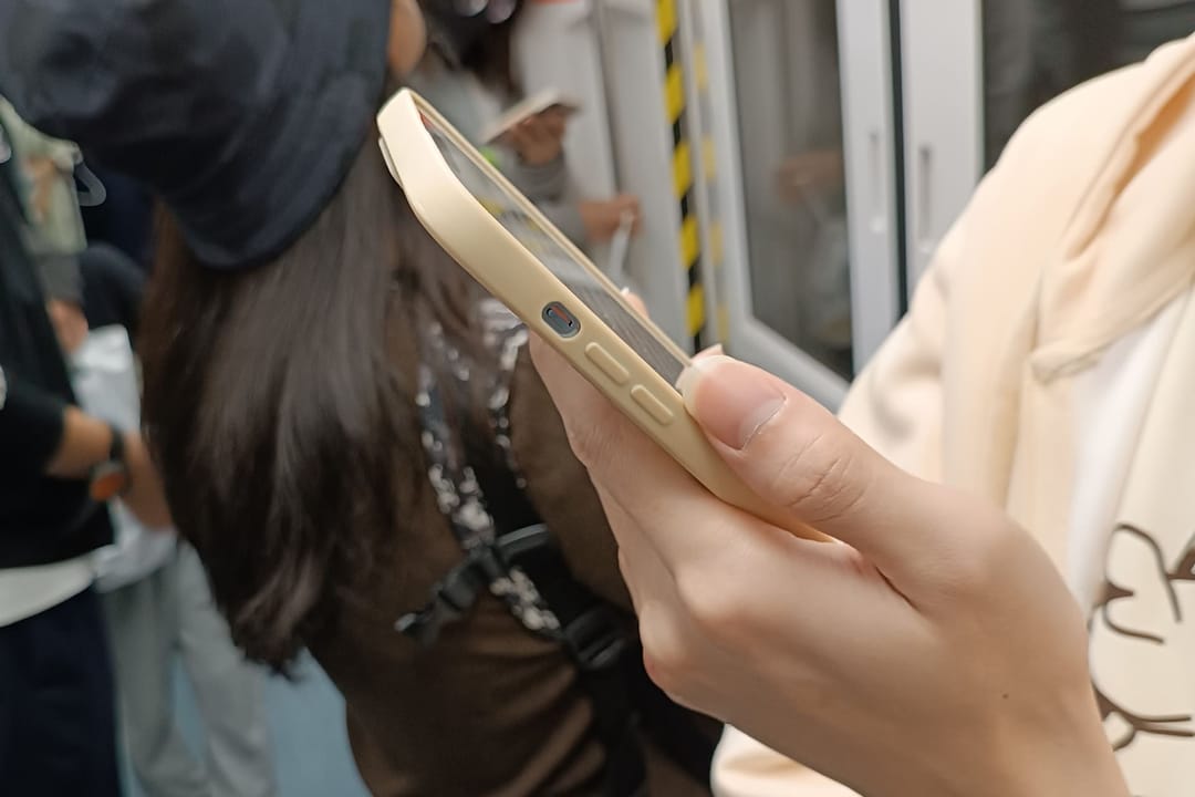 Une main tient un smartphone dans une rame de métro à Shenzhen, écran vu par la tranche, d'autres passagers debout autour et portes de la rame en arrière-plan