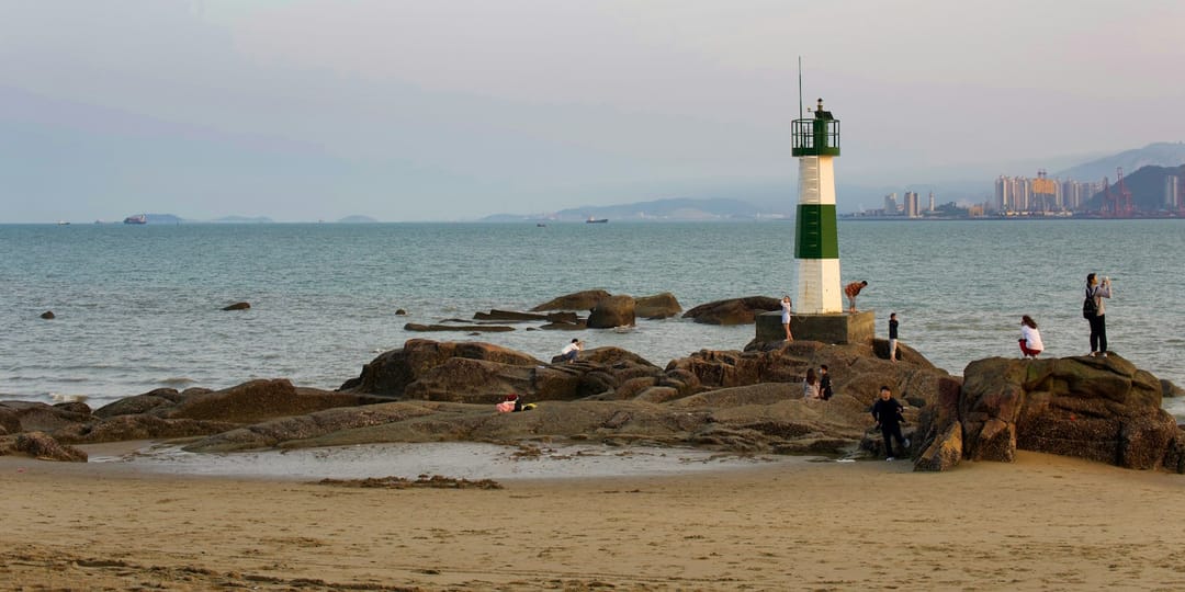 Une plage de sable sur l'île de Gulangyu en fin de journée : au premier plan des rochers bruns émergeant du sable, au milieu un petit phare rayé vert et blanc sur un socle de pierre entouré de promeneurs, et de l'autre côté de l'eau la silhouette du port de Xiamen avec ses grues et ses tours dans la brume