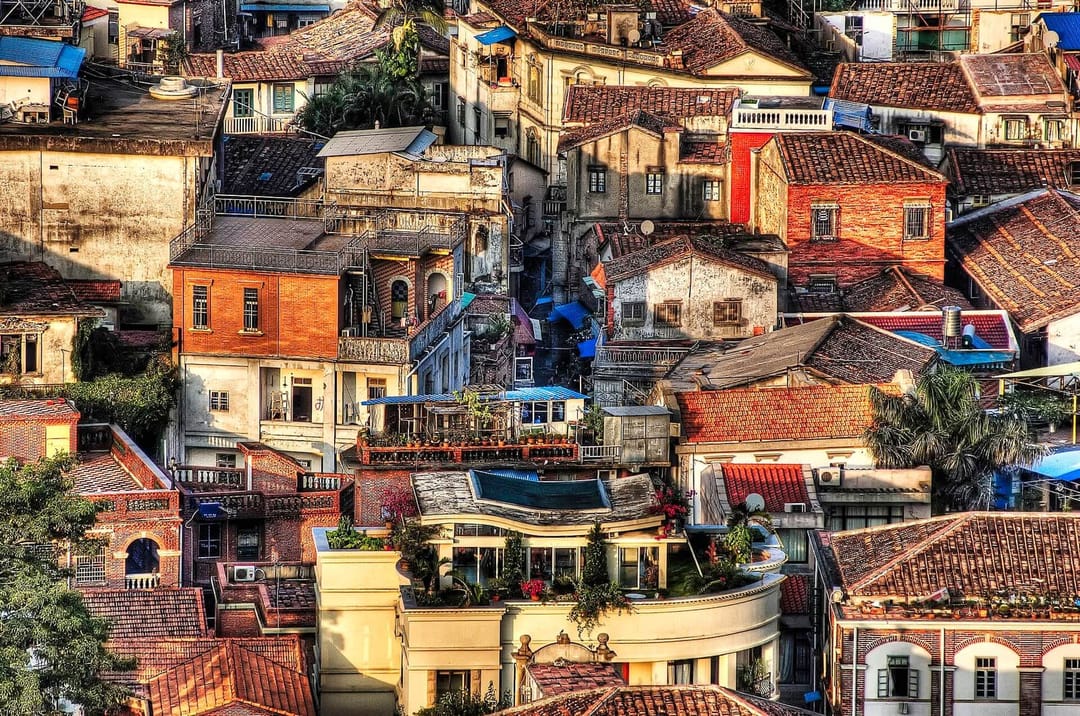 Les toits de Gulangyu vus d'en haut : maisons de brique rouge, villas crépies à balustrades, un immeuble aux courbes arrondies et des terrasses plantées, imbriqués les uns dans les autres sans qu'une rue soit visible entre eux