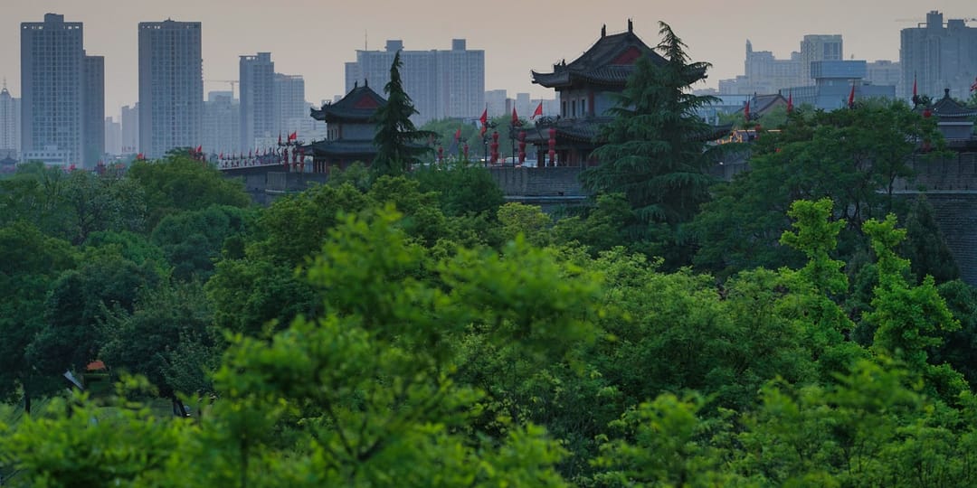 Le rempart de Xi'an au crépuscule : une tour de guet à toit recourbé et lanternes rouges émerge d'une masse d'arbres verts, une rangée de drapeaux rouges longe le chemin de ronde, et derrière se dressent les tours d'habitation de la ville moderne dans une brume orangée