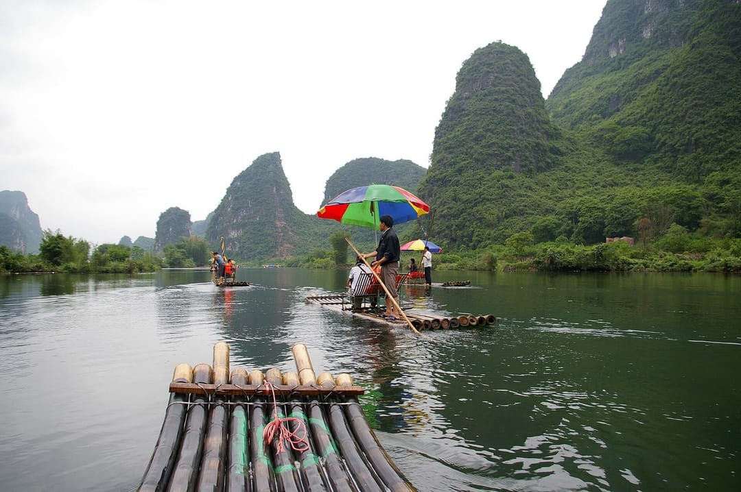 Descente en radeau de bambou sur la rivière Yulong, vue depuis l'avant d'un radeau : le perchiste debout à l'arrière du radeau qui précède, sous un parasol multicolore, deux autres radeaux plus loin sur l'eau, et les pics calcaires couverts de végétation sur les deux rives