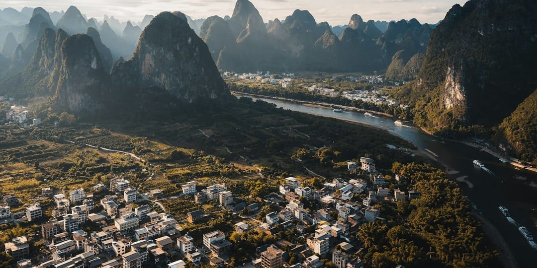 Vue aérienne d'un bourg de la vallée de Yangshuo au lever du jour : des immeubles de quatre à six étages serrés au pied des pics calcaires, les champs et les vergers qui reprennent dès la dernière maison, et la rivière large avec ses bateaux de croisière sur la droite
