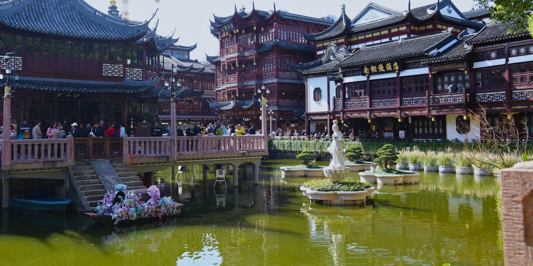 Le bazar Yuyuan vu depuis la rive de l'étang : pavillons de bois rouge sombre à toits de tuiles recourbées, une maison de thé sur pilotis à gauche, un pont de pierre couvert de visiteurs, et au premier plan l'eau verte où flottent des îlots plantés de bonsaïs autour d'une statue blanche