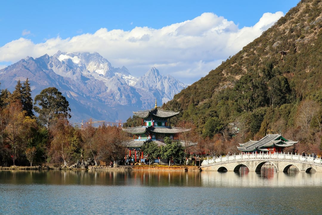 L'étang du Dragon Noir à Lijiang, un pavillon traditionnel et un pont de pierre en arche sur l'eau calme, la montagne enneigée du Dragon de Jade en arrière-plan sous un ciel bleu